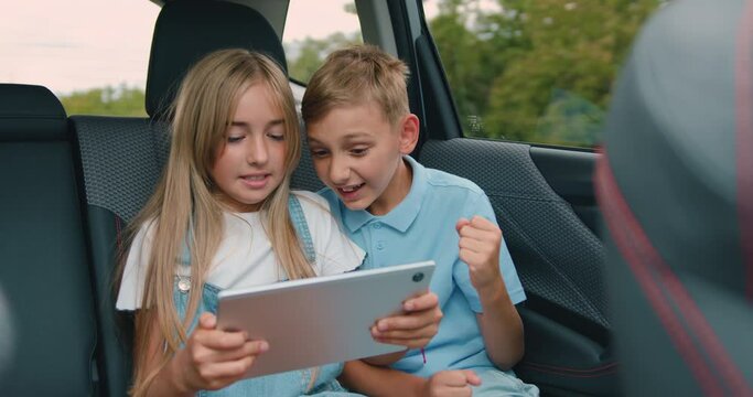 Children boy and girl sit in car using digital tablet for games and entertainment, celebrate vin and give high fives traveling by car. Concept of a family holiday.