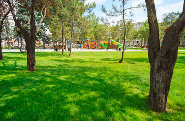 city park in day, beautiful nature, trees and lawn, children's playground with children playing in the distance, late summer and early autumn season, urban architecture, street and random people