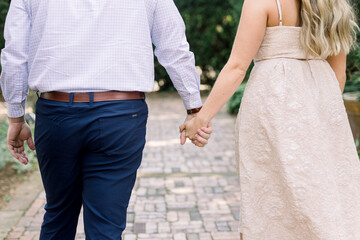 An engaged couple are holding hands and walking away. She is wearing a cream dress, and he has a dress shirt, belt, and pants on