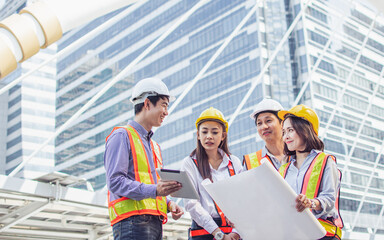 Group of asian adult civil engineers wearing safety helmet, discussing, holding papers of building construction project plan while standing outdoor site.