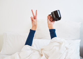 Close up of hands making peace sign with coffee mug peaking from duvet