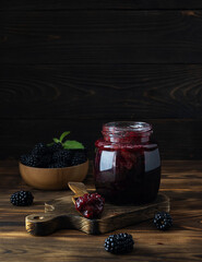Blackberry jam in a transparent jar on a wooden board. Blackberries in a wooden bowl in the background. Wooden spoon with jam on the table. Dark wooden background