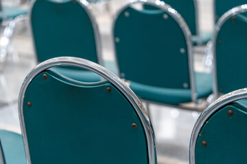 Many empty green leather chairs in a wide hall room, lights on. A floor reflect the objects. Give a feeling of emptiness.