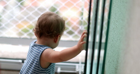 Cute baby standing by home window during lockdown in apartment