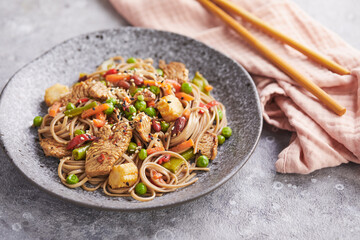 Wok with turkey meat, soba noodles, corn, green peas, green beans and carrots served on gray background with chopsticks. Asian food, concept of street food