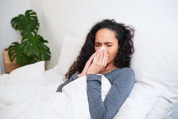A young multi-ethnic woman blows her nose while recovering from flu in bed