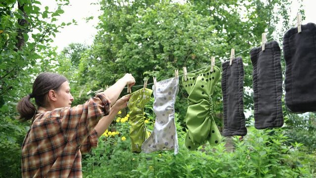 Middle age woman hanging reusable diapers to dry on clothesline in garden. Modern eco friendly cloth nappy for infant child hygiene. Sustainable lifestyle, zero waste concept.