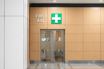 First Aid room inside a building. Decorated with pain light brown wall, push and pull door at an entrance and left open door to a scary walk way at a background with dim light.