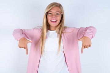 caucasian blonde little girl wearing pink jacket and glasses over white background with positive expression, points down with both index fingers, has broad interested smile. Look there, please.