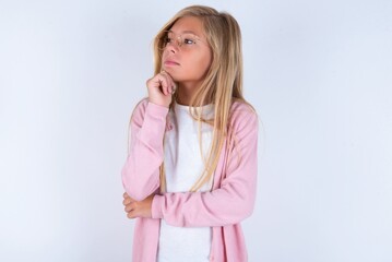 Portrait of thoughtful caucasian blonde little girl wearing pink jacket and glasses keeps hand under chin, looks away trying to remember something or listens something with interest. Youth concept.