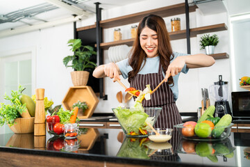 Happy Asian woman wearing apron, tossing the vegetarian salad with wooden spatulas. Preparing a healthy salad with fresh vegetables such as carrot, tomato cabbage and green oak in the home kitchen.