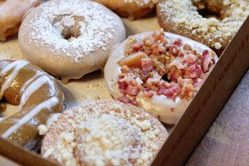 Picture of assorted donuts in a box with chocolate frosted, powdered and bacon sprinkled donuts.