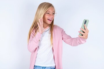 Portrait of happy friendly caucasian blonde little girl wearing pink jacket and glasses over white background taking selfie and waving hand, communicating on video call, online chatting.