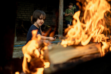 The boy is sitting near the fire and burning sticks. The child spends time outdoors in the summer