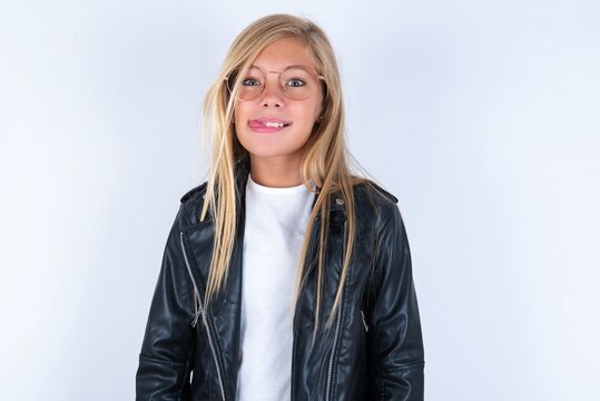 Funny beautiful caucasian blonde little girl wearing biker jacket and glasses over white background makes grimace and crosses eyes plays fool has fun alone sticks out tongue.