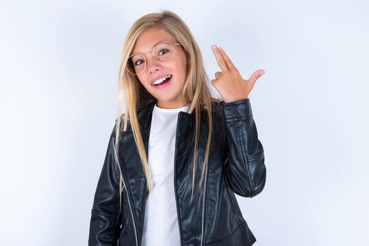 beautiful caucasian blonde little girl wearing biker jacket and glasses over white wall foolishness around shoots in temple with fingers makes suicide gesture. Funny model makes finger gun pistol