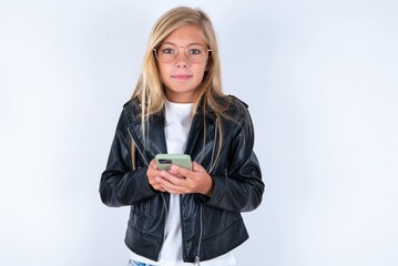 Fototapeta premium Portrait of serious confident beautiful caucasian blonde little girl wearing biker jacket and glasses over white background holding phone in two hands
