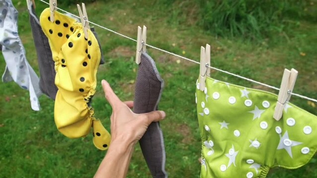 Close up person hand removes dried reusable diapers from clothesline in garden. Modern eco friendly cloth nappy for infant child hygiene. Sustainable lifestyle, zero waste concept. Slow motion.