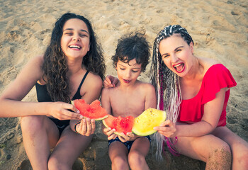 family, leisure and people concept - happy mother and two children having picnic on summer beach and eating red and yellow watermelon