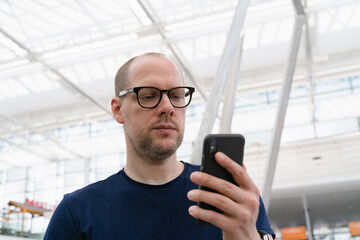Middle-aged business man with glasses in casual T-Shirt attire holding mobile cell phone in his hand checking messages and emails on his smartphone at bright airport terminal ahead of travel departure - 530873334