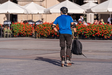 A young man in a helmet on roller skates with a backpack in his hand on a city street on a sunny day against the background of summer restaurant umbrellas. Concepts of youth, sport, modern lifestyle