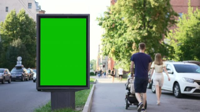 4k Mockup billboard stands and people walk in city street spbd. Green empty poster is standing and pedestrians walking in modern town on summer day. There is vertical screen designed for advertising