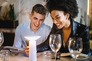 Multiethnic couple sitting at restaurant table and watching funny content on the smart phone screen