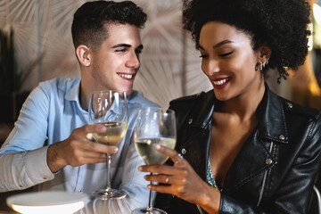 Multiracial romantic couple sitting on the restaurant table celebrate with a toast with white wine