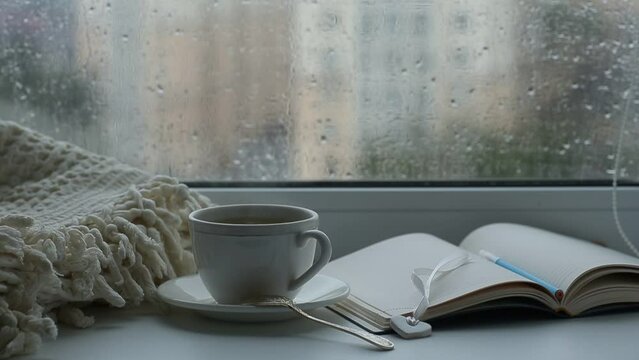 Rain outside the window, a cup of tea, a blanket and a notebook on the windowsill, drops flow down the glass.
