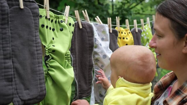 Mother with little baby hang reusable diapers to dry on clothesline in the garden. Modern eco friendly cloth nappy for infant child hygiene. Sustainable lifestyle, zero waste concept.