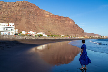 Woman in dress walking on the beach Playa Valle Gran Rey during sunset seen from Promenade de Playa de La Calera in Valle Gran Rey on La Gomera, Canary Islands, Spain, Europe. Dark volcanic sand beach