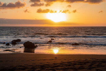 Scenic view during sunset on the volcanic sand beach Playa del Ingles in Valle Gran Rey, La Gomera, Canary Islands, Spain, Europe. Sun beam reflection on water surface. Stones in the foreground