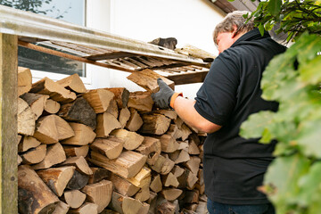 Ein 50 jähriger Mann hat eine Lieferung Brennholz bekommen und räumt die Buchen Scheite in das Kaminholzregal. Draußen vor dem Haus ist das Regal mit einem Dach gegen Witterungseinflüsse ausgestattet.