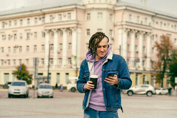 Fototapeta premium Hipster man with dreadlocks drinking hot coffee on street together with cell phone Outdoors city background. Young male in warm clothes drinking hot beverage