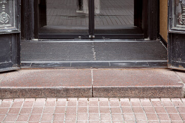granite threshold at entrance to open iron doors through foot mat to glass door with black frame, central entrance to store from pedestrian sidewalk with stone tiles, close-up architecture.