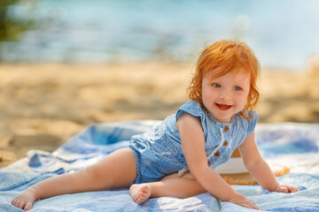 Red hair baby girl in blue clothes is resting on the sand on the beach in summer. 