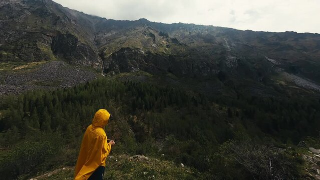 beginnig the flight by fpv race drone towards the mountain past the drone pilot in yellow hooded raincoat in vr goggles standing on the bloomy hillside in highlands over the mountain river