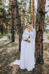 A beautiful brunette bride in a white long dress with a bouquet of roses stands in a coniferous forest in nature near a tree. Wedding portrait, photography.