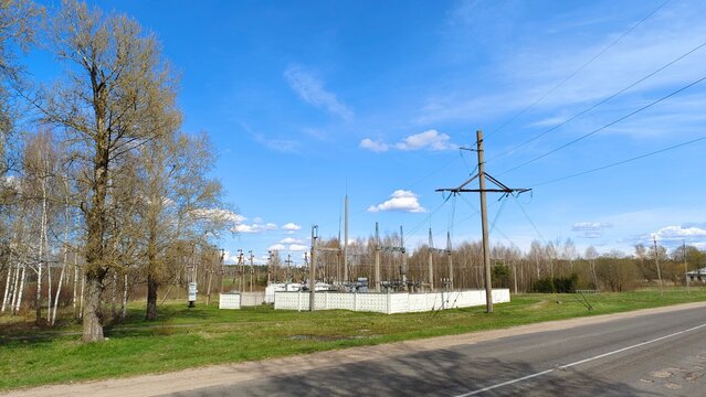 A transformer station with concrete poles, ceramic insulators, and lightning rods is located near the road on a green lawn and surrounded by a fence. A forest grows behind the station. Sunny