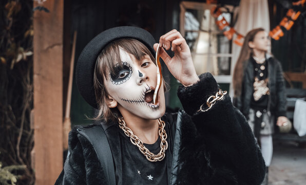 Beautiful scary little girl celebrating halloween, chewing gummy worm. Terrifying face makeup and witch costume, stylish image. Horror, fun at children's party in barn on street. Hat, fur coat, chain