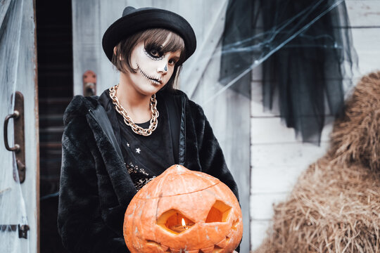 Beautiful scary little girl celebrating halloween. Terrifying black, white half-face makeup,witch costume, stylish image. Fun at children's party in barn on street. Hat,jacket, pumpkin jack-o-lantern
