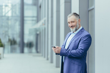 Senior handsome gray-haired man stands in a suit against the background of a modern building, uses the phone, checks calls, mail, messages. He looks at the camera, smiles.
