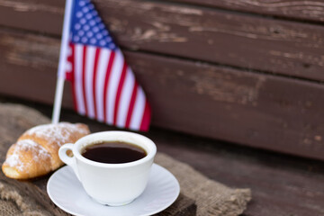 A cup of coffee and a croissant on a wooden table against the background of the American flag