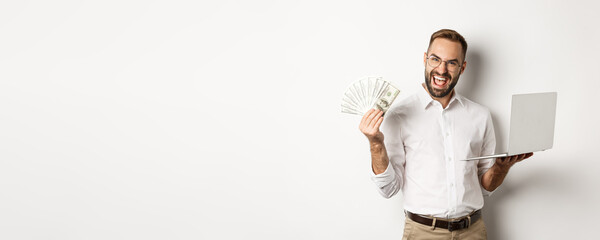 Business and e-commerce. Excited businessman holding money dollars and laptop, working online,...