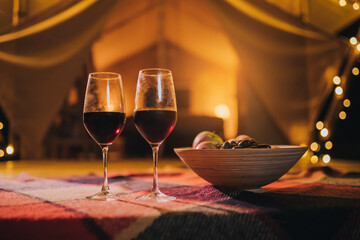 Two glass of red wine and plate with fruits standing on background of cozy glamping tent on autumn evening. Luxury camping tent for outdoor holiday and vacation. Lifestyle concept - 530909147