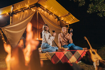 Two Smiling female friends drinking wine and eating fruits sitting in cozy glamping tent in autumn evening bonfire. Luxury camping tent for outdoor holiday and vacation. Lifestyle concept