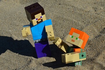 Fototapeta premium LEGO Minecraft action figure of Steve in blue shirt helping his female friend Alex from shifty sand pit on a beach. Summer afternoon sunshine. 