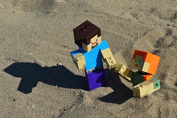 Fototapeta premium LEGO Minecraft large action figure of Steve in blue shirt pulling out of desert shifty sand pit his female friend Alex. Summer afternoon sunshine. 