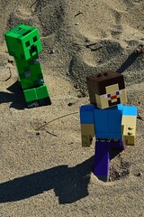 Fototapeta premium LEGO Minecraft large figure of main character Steve chased by green explosive Creeper mob on sandy dunes, summer afternoon sunshine.