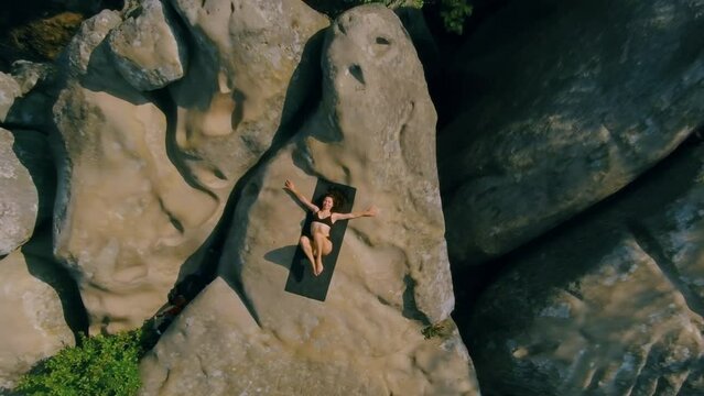 Woman lying on mat, twisting pelvis with her waist. top view. Beautiful sports lady training at dawn on rocky terrain. Pretty fitness girl in sportswear during workout session on rocky outside. Aerial